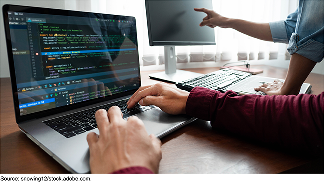 Photo showing two people working side by side. We only see their hands. One is typing on a laptop. The other is pointing to a desktop computer screen.