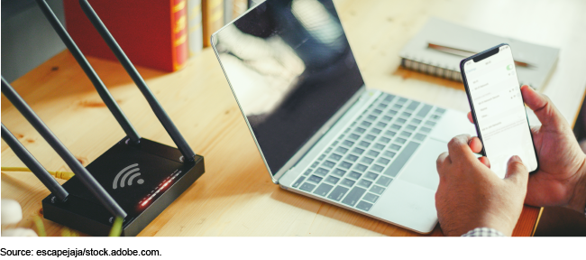 A router and a laptop on a desk with the hands of someone holding a smartphone in front of the laptop.