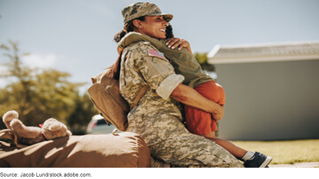 A woman wearing military fatigues hugs a small child while kneeling on the ground.