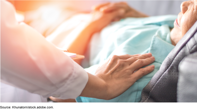 An image of an elderly person in a bed with a person's hand outstretched on the person's shoulder.