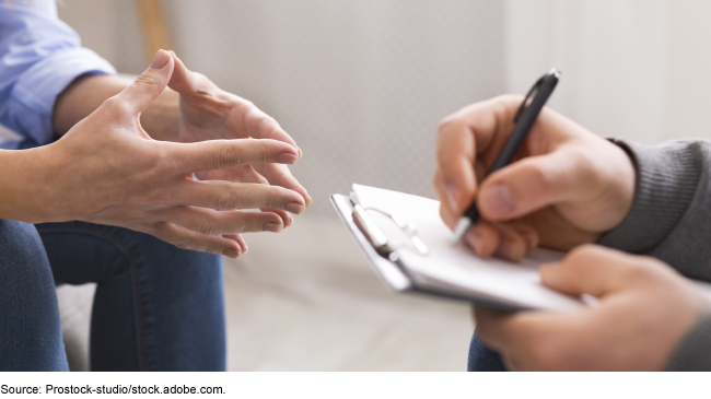 A person is holding a pen and clipboard sitting across from another person.