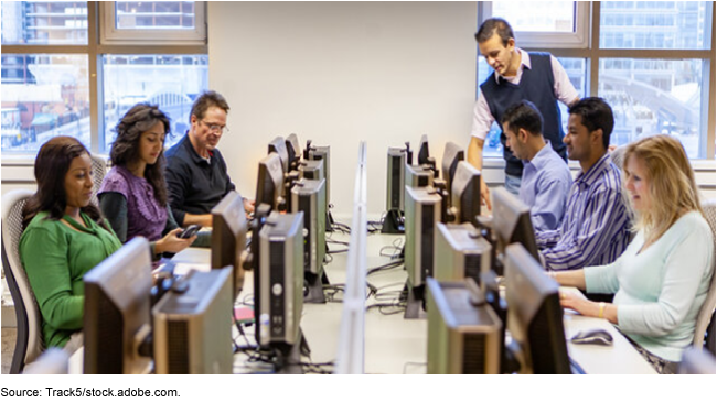 Six adults use computers in a training class as they are assisted by an instructor.