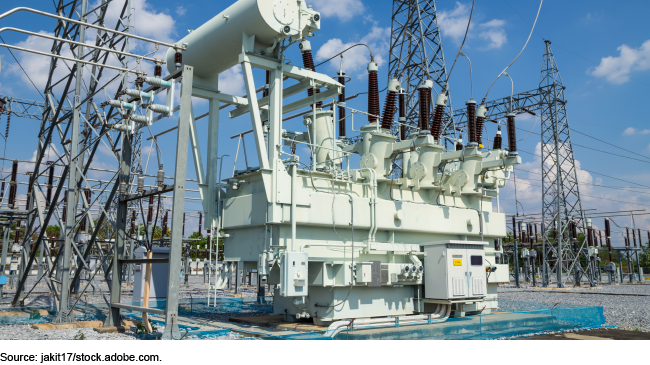 Photo showing a large power transformer. It's a big metal box with large cables attached and power lines in the background.