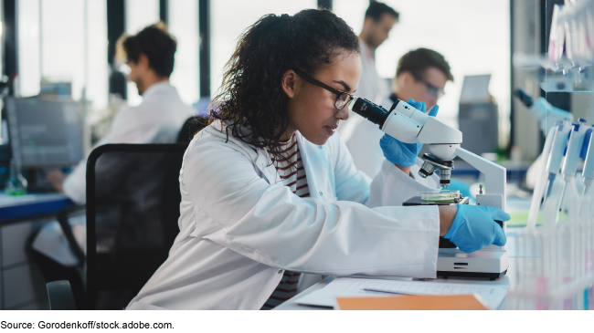 Stock image showing a woman in a lab coat and gloves looking through a microscope. 