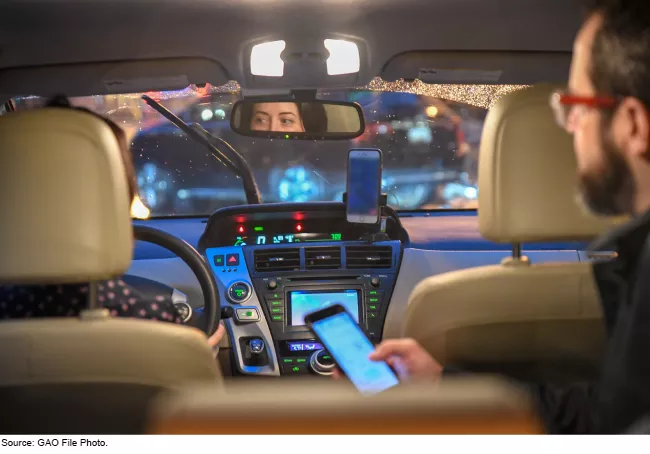 Image showing the inside of a car with one person behind the wheel and another in the backseat holding a phone.