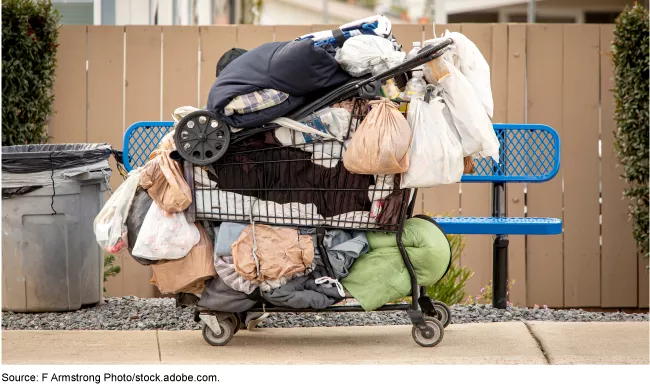 Stock image showing a shopping cart next to a park bench. The shopping cart is overloaded with belongings.