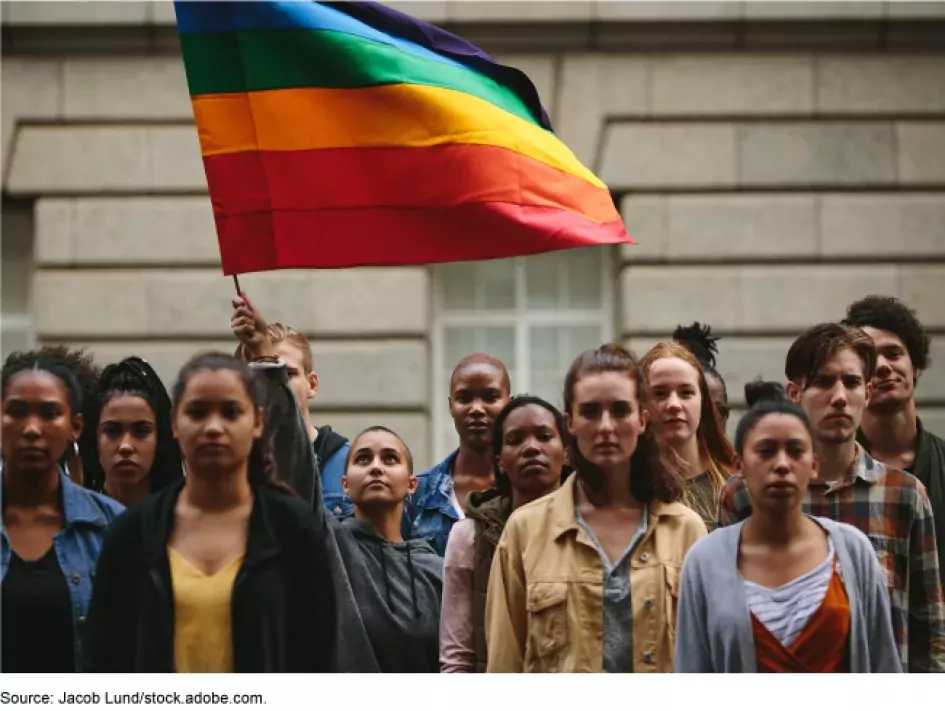 Photo of young people standing with a Pride flag outside of what looks like a municipal building. 