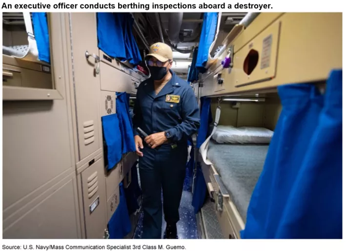 Photo showing a Navy officer walking down a hallway that has bunk beds installed on either side with little curtains.